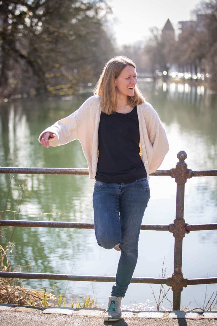 ein Foto von Sandra. Sie steht an einer Brücke, dahinter ist ein Fluss. Die Sonne scheint. Sandra lacht und schaut zur Seite.