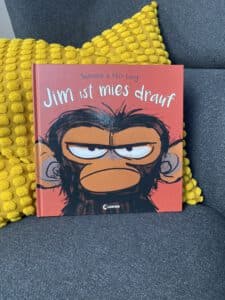ein Foto. Man sieht das Buch auf einem grauen Sessel. Da ist im Hintergrund ein gelbes Kissen. Das Buch ist rot. Da ist ein Kopf von einem Schimpansen drauf gemalt. Der Schimpanse schaut sehr grimmig. Über dem Schimpansen steht in weiß der Titel von dem Buch: "Jim ist mies drauf." Man erkennt, dass das ganze Bild witzig gemeint ist, obwohl der Schimpanse so böse schaut.